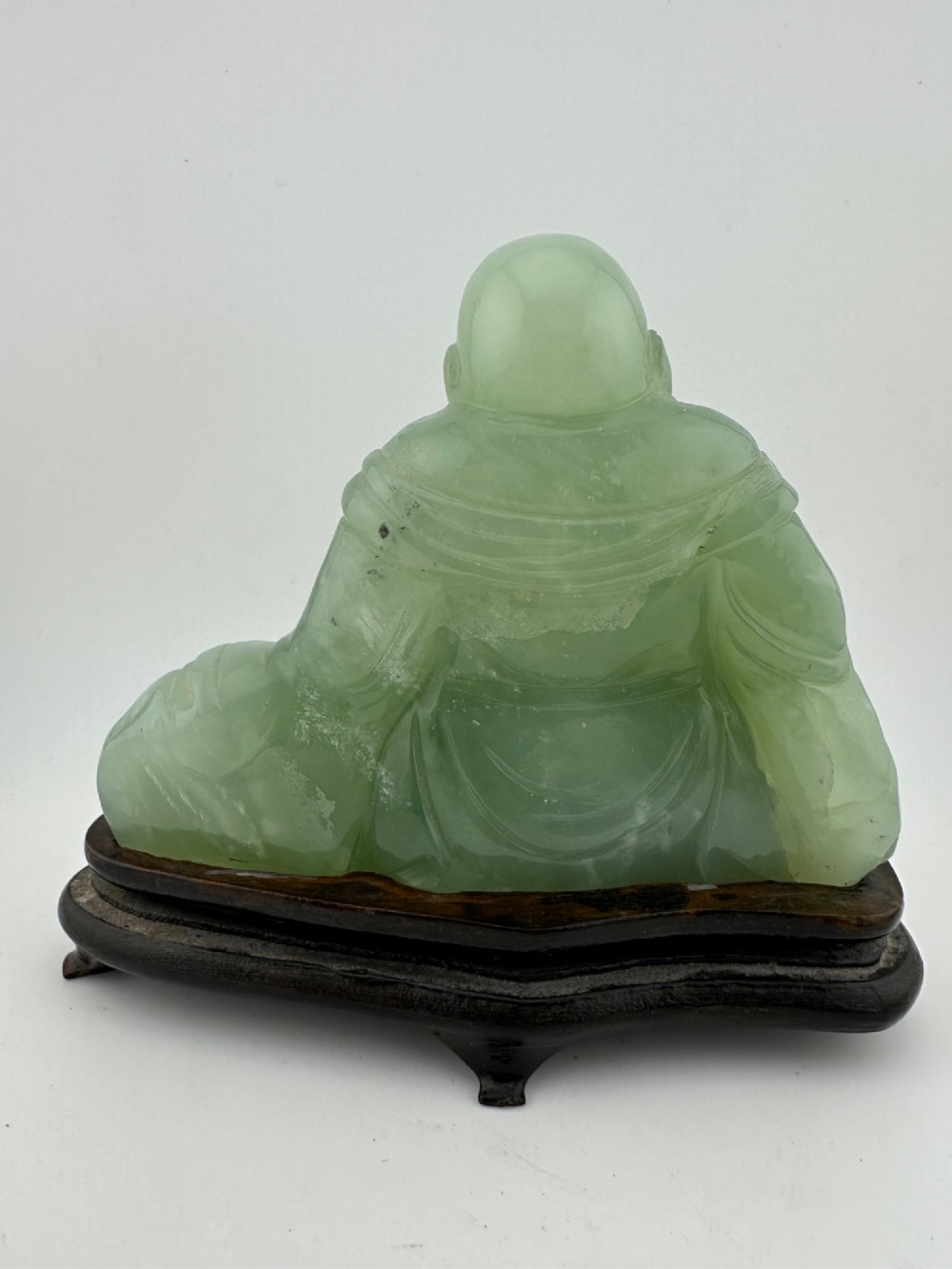 A hand-carved light green jadeite sculpture depicting a seated Laughing Buddha resting on a dark wooden fitted base. View V