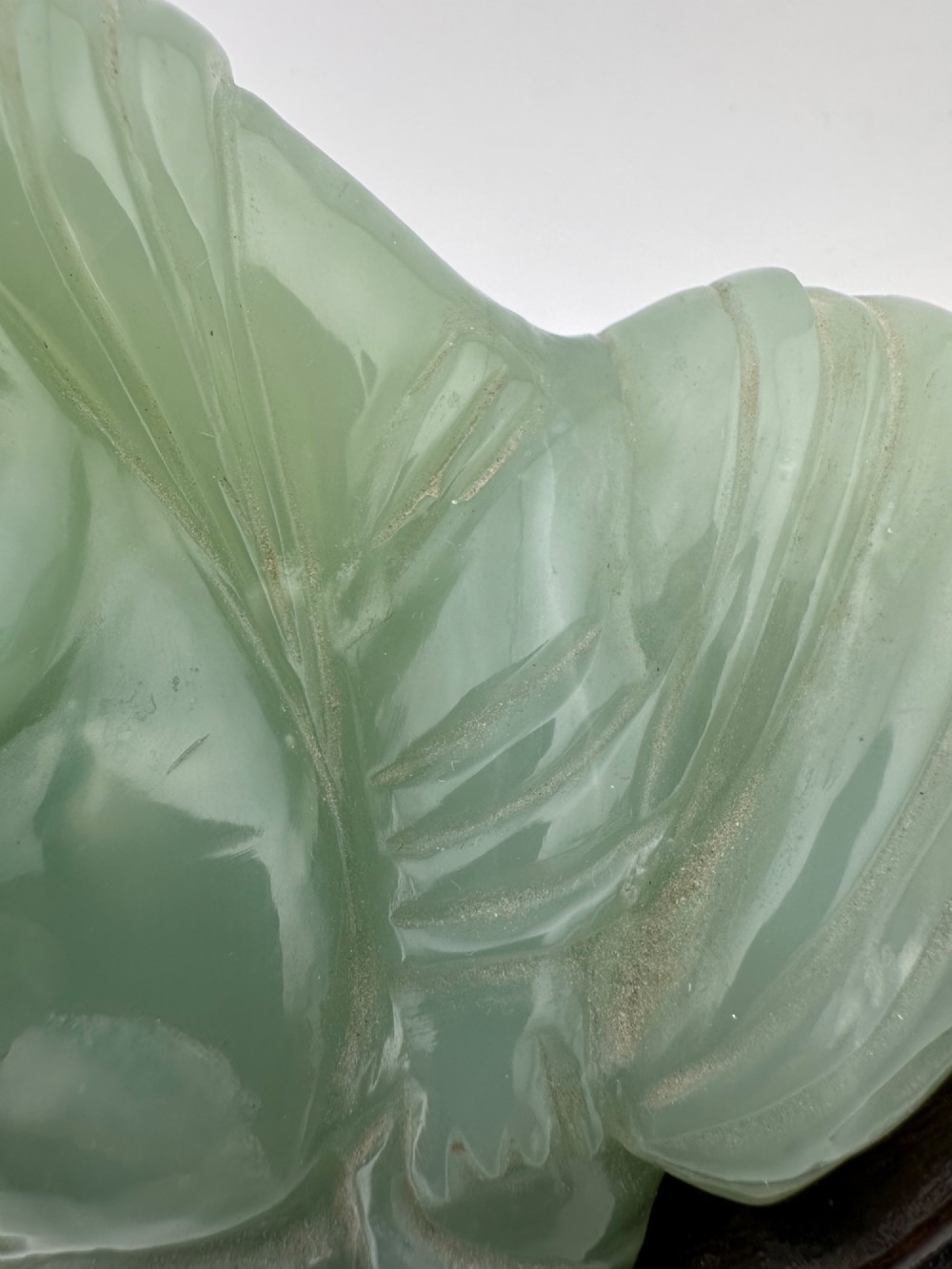 A hand-carved light green jadeite sculpture depicting a seated Laughing Buddha resting on a dark wooden fitted base. View XII
