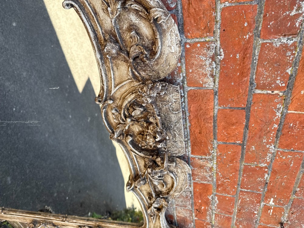 A large nineteenth-century overmantel mirror featuring a distressed cream painted wood and gesso frame decorated with intricate Rococo scrollwork and a central crest. View VII