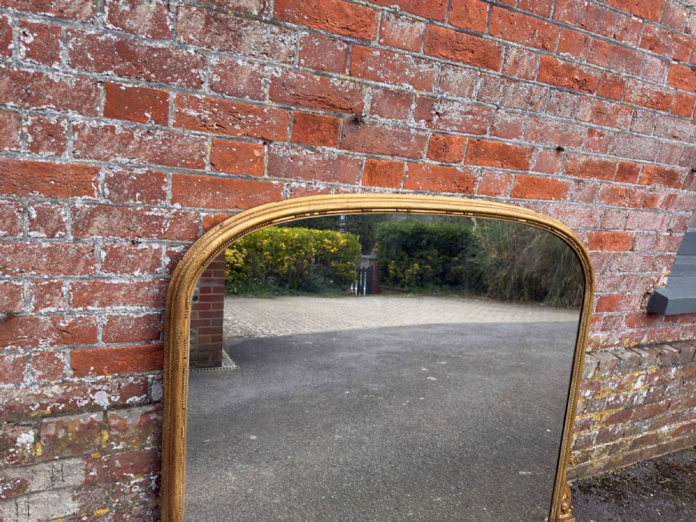 A large 19th-century English giltwood overmantel mirror featuring an arched top frame decorated with beaded moulding and carved scrollwork details at the base. View IV