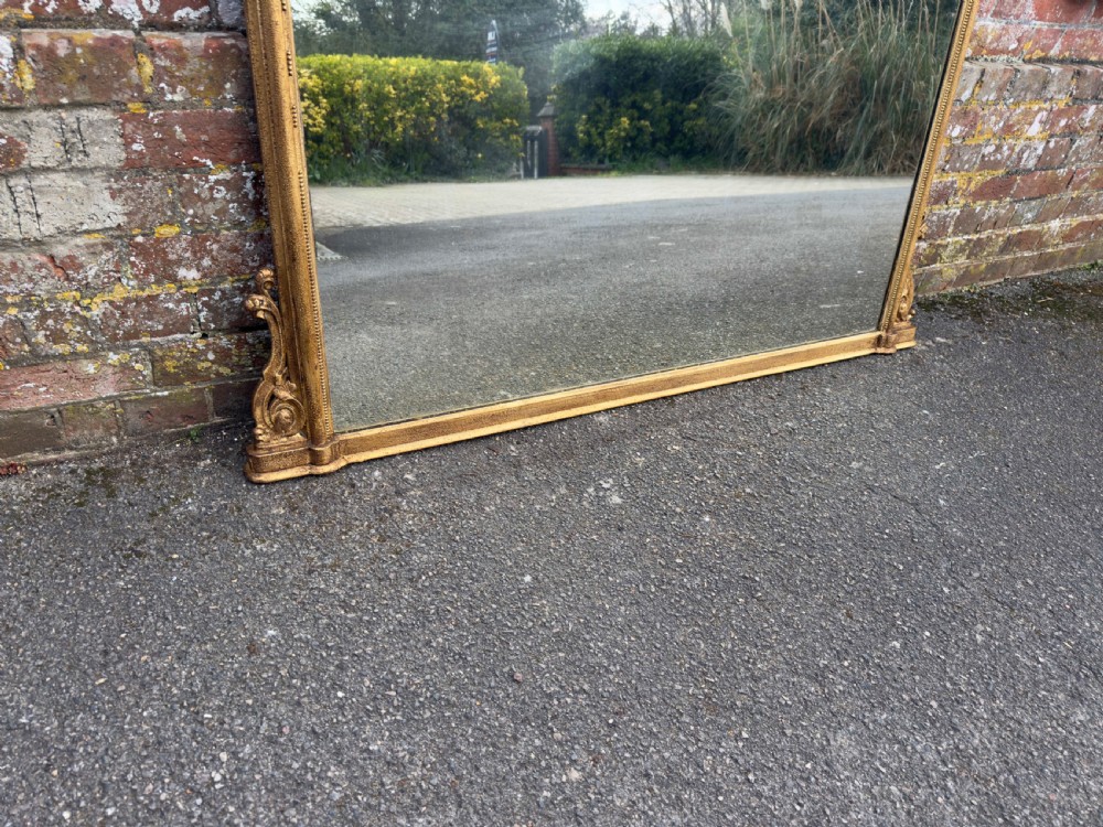 A large 19th-century English giltwood overmantel mirror featuring an arched top frame decorated with beaded moulding and carved scrollwork details at the base. View V