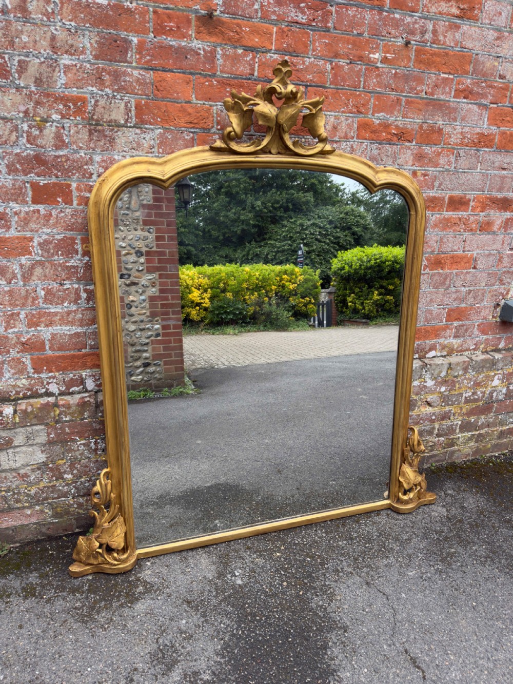 A large 19th-century gilded overmantel mirror featuring a shaped upper frame with a carved floral crest and decorative scroll accents at the bottom corners.