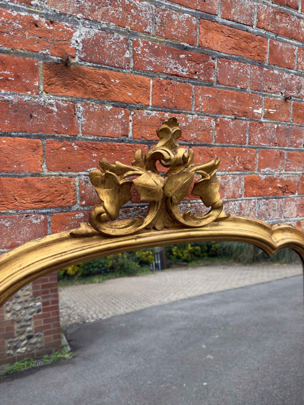 A large 19th-century gilded overmantel mirror featuring a shaped upper frame with a carved floral crest and decorative scroll accents at the bottom corners. View II