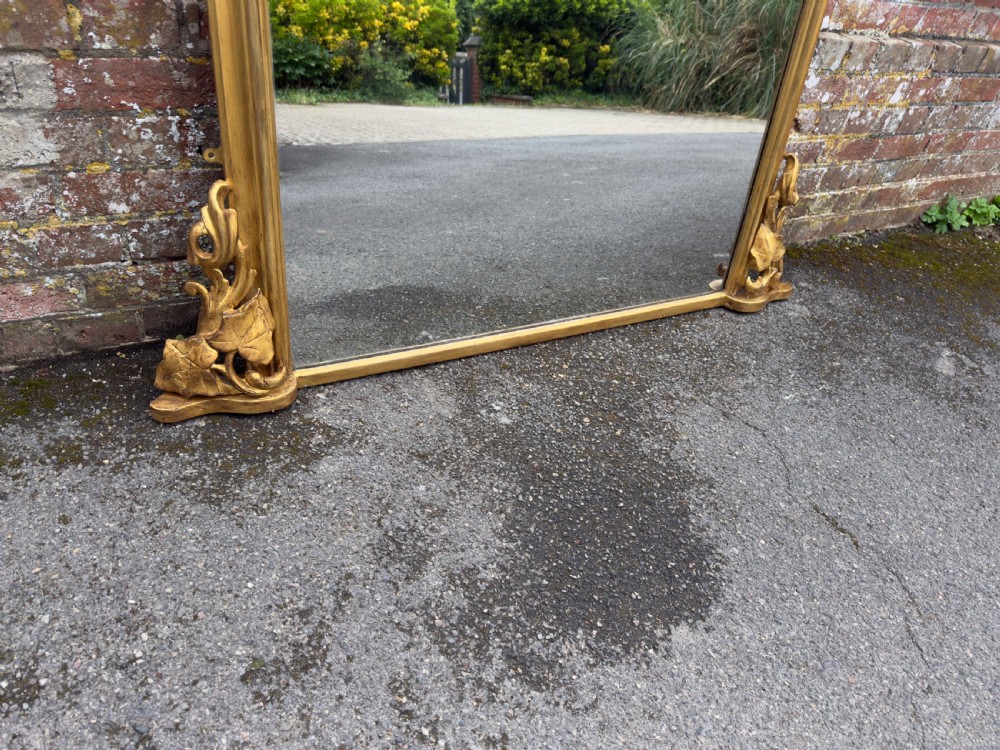 A large 19th-century gilded overmantel mirror featuring a shaped upper frame with a carved floral crest and decorative scroll accents at the bottom corners. View V