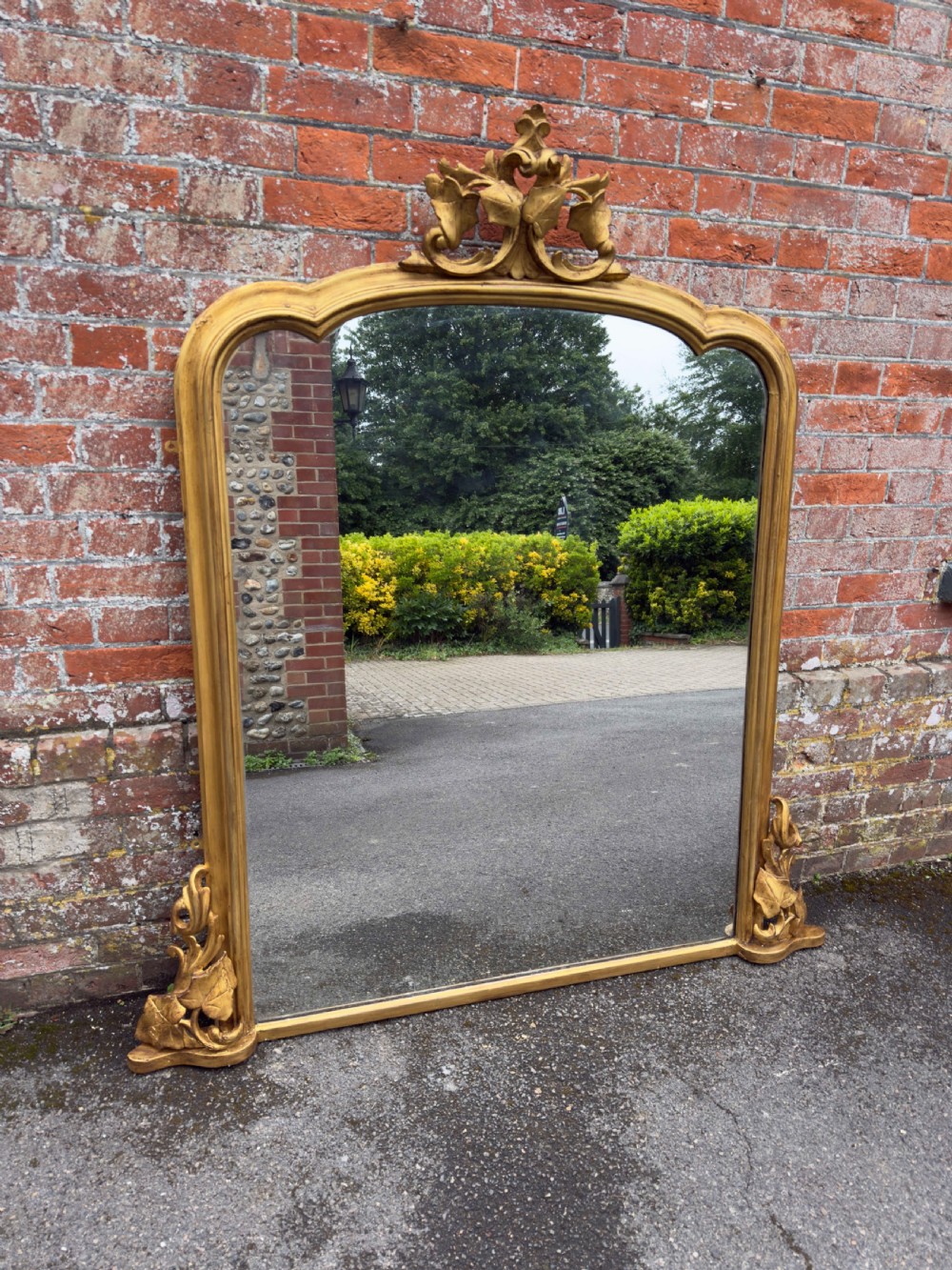 A large 19th-century gilded overmantel mirror featuring a shaped upper frame with a carved floral crest and decorative scroll accents at the bottom corners. View VIII