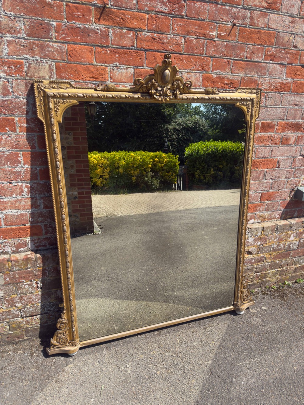 A large 19th-century English giltwood and gesso overmantel mirror featuring an ornate carved central crest, moulded frame, and scrolled side feet.