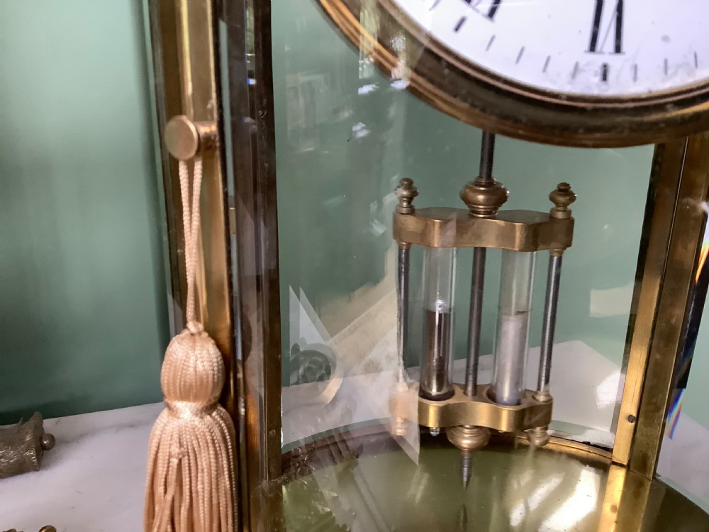 An oval brass and glass mantel clock with a white enamel dial showing Roman numerals and a visible mercury pendulum inside. View II
