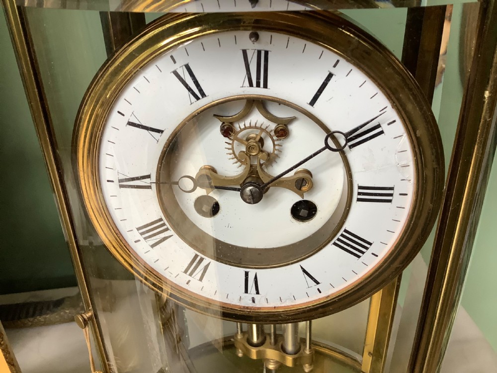 An oval brass and glass mantel clock with a white enamel dial showing Roman numerals and a visible mercury pendulum inside. View III