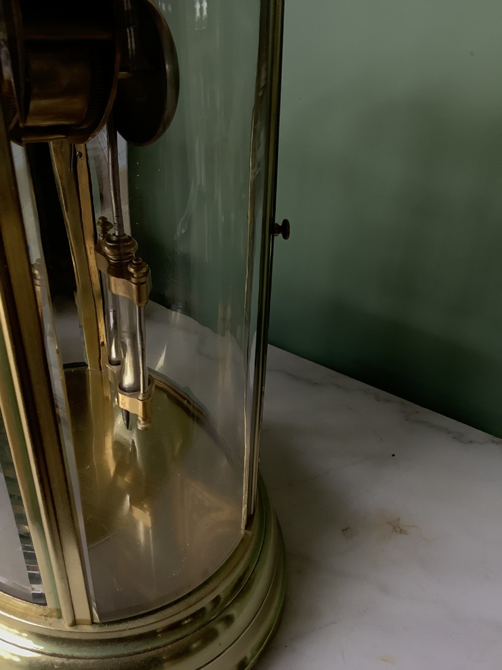 An oval brass and glass mantel clock with a white enamel dial showing Roman numerals and a visible mercury pendulum inside. View V