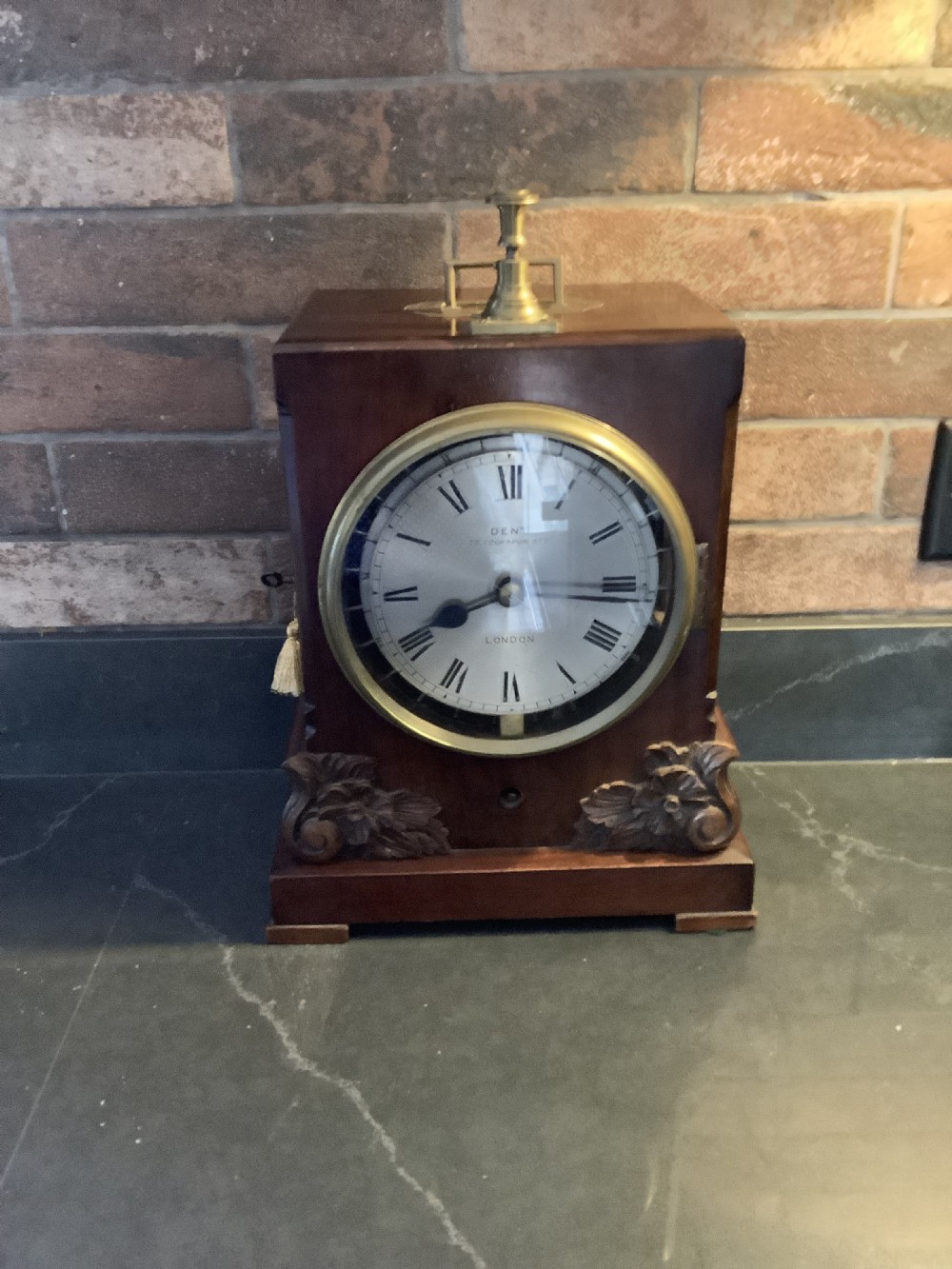A Victorian mahogany night watchman clock featuring a circular dial with Roman numerals, brass bezel, top mechanism, and carved foliate details at the base.