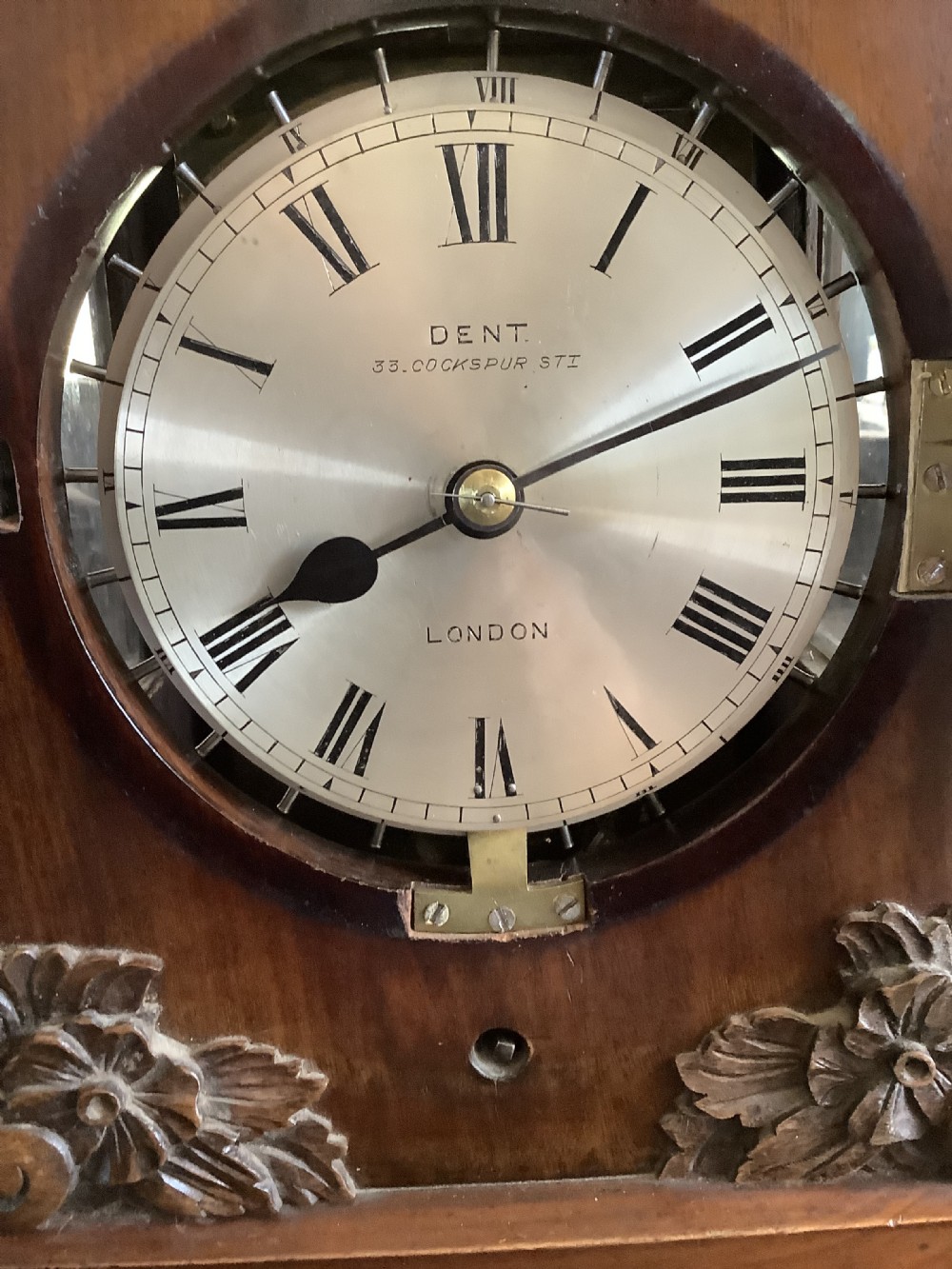 A Victorian mahogany night watchman clock featuring a circular dial with Roman numerals, brass bezel, top mechanism, and carved foliate details at the base. View II