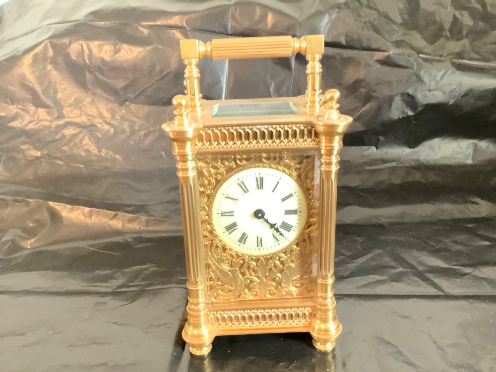 An ornate nineteenth-century French gilt brass carriage clock featuring fluted corner columns, intricate leaf scrollwork, a white enamel dial with Roman numerals, and a hinged top handle. View II