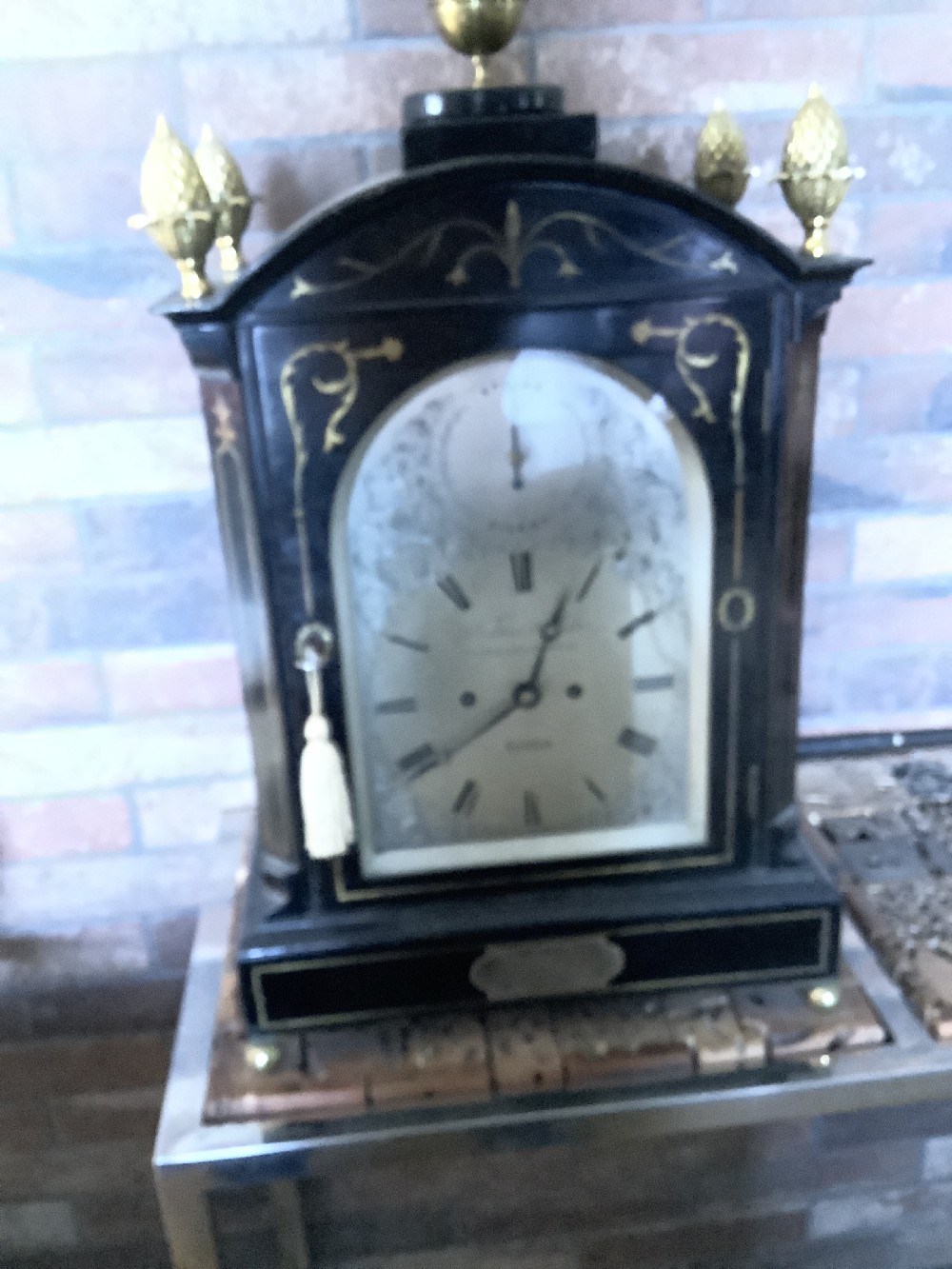 An antique arched ebonised wood bracket clock featuring brass pineapple finials, brass inlay, and an engraved metal dial with Roman numerals. View XII