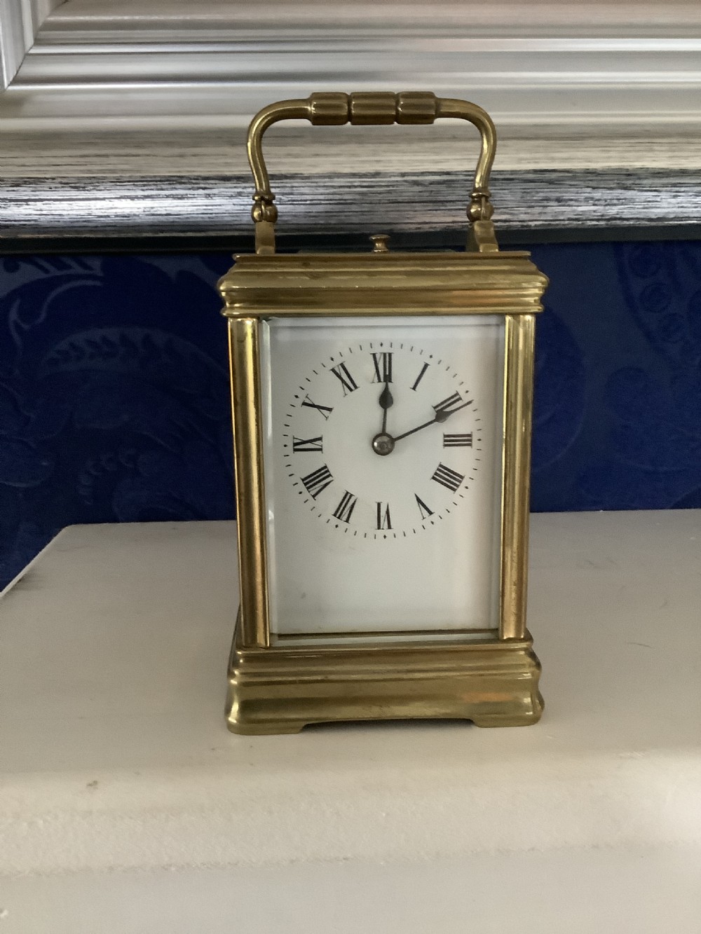 A miniature brass carriage clock featuring a top carrying handle, glass side panels, and a white enamel dial with black Roman numerals.