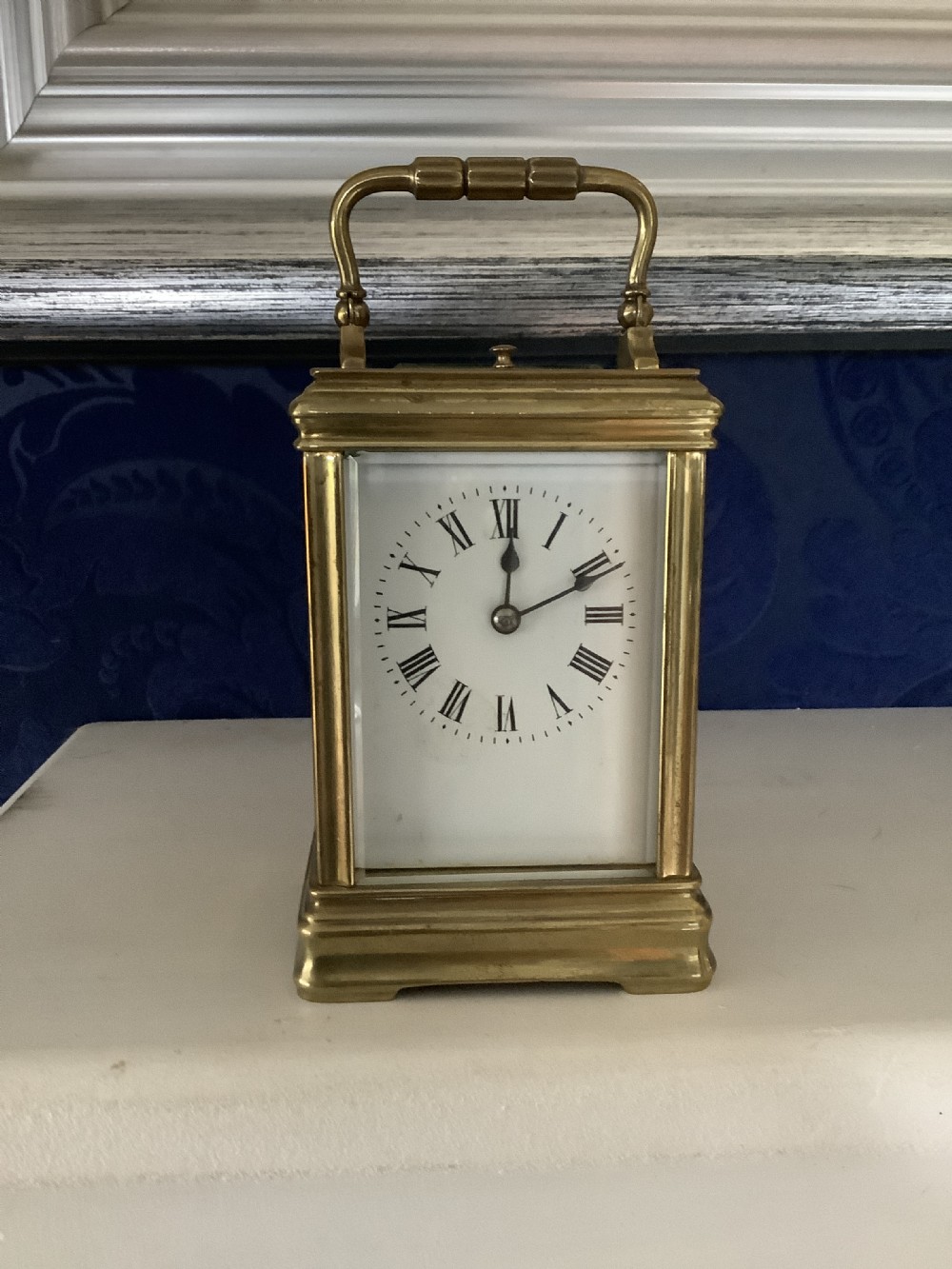 A miniature brass carriage clock featuring a top carrying handle, glass side panels, and a white enamel dial with black Roman numerals. View II