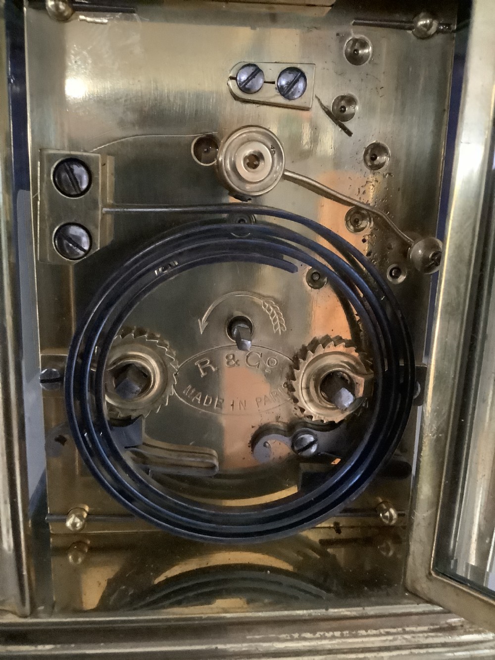 A miniature brass carriage clock featuring a top carrying handle, glass side panels, and a white enamel dial with black Roman numerals. View III