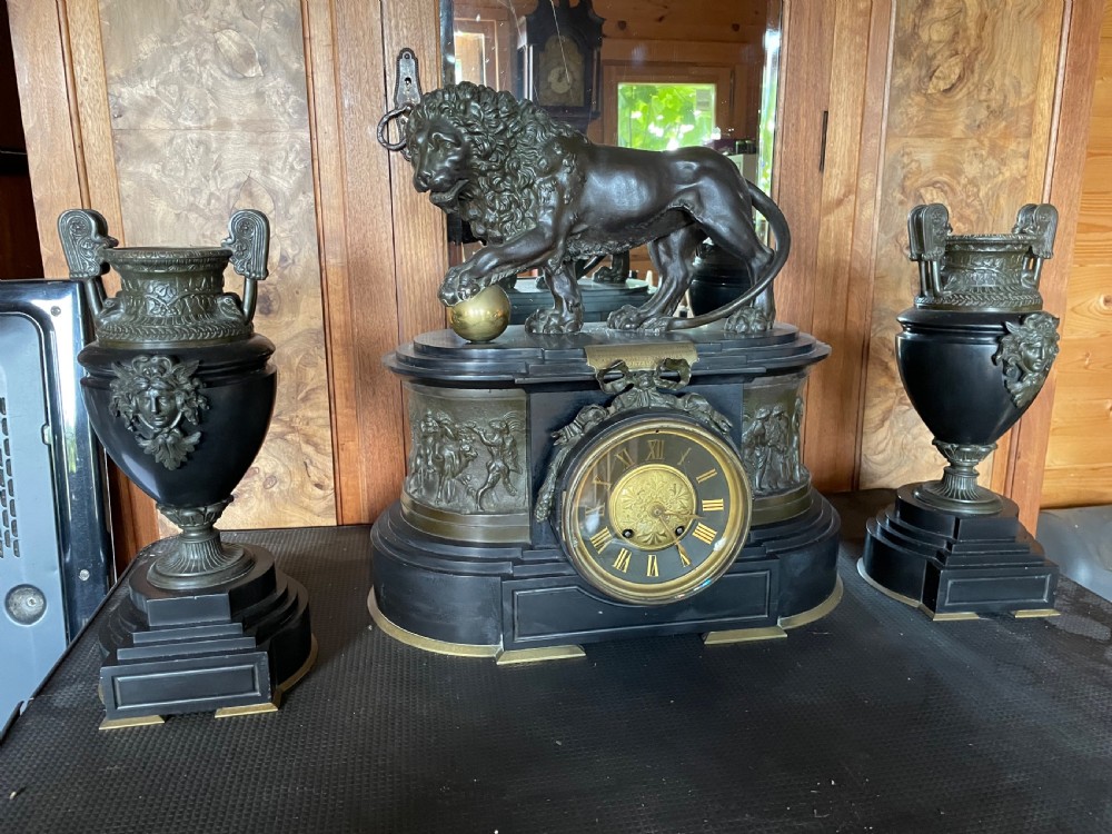 An antique French mantel clock garniture set featuring a black stone clock topped with a bronze lion alongside two matching classical urns.