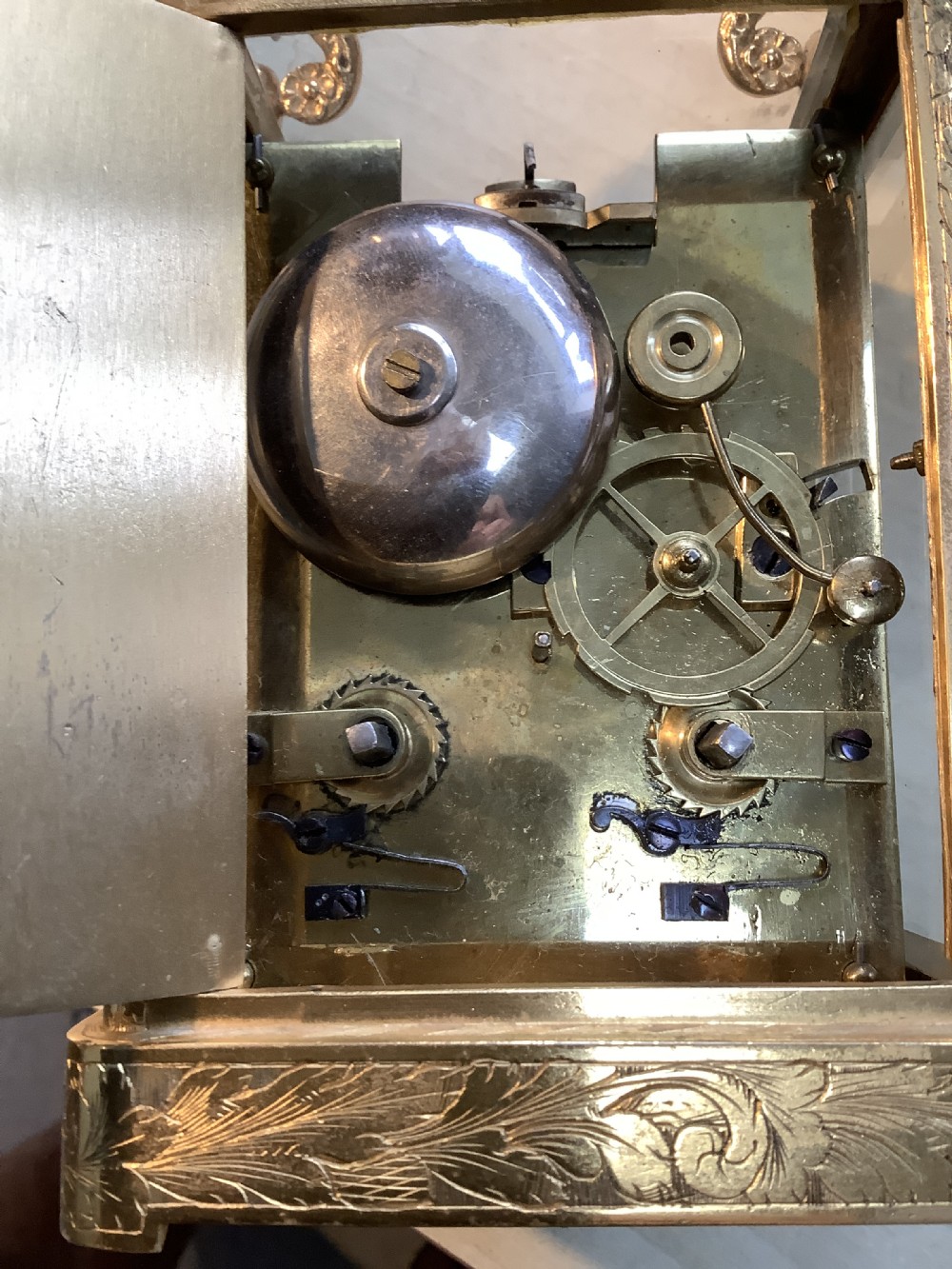A rectangular gilt brass French carriage clock featuring ornate floral scroll engravings, glass side panels, a decorative carrying handle, and a white enamel dial. View II