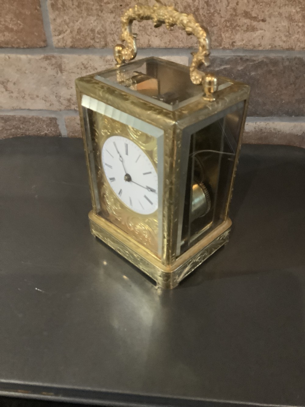 A rectangular gilt brass French carriage clock featuring ornate floral scroll engravings, glass side panels, a decorative carrying handle, and a white enamel dial. View III