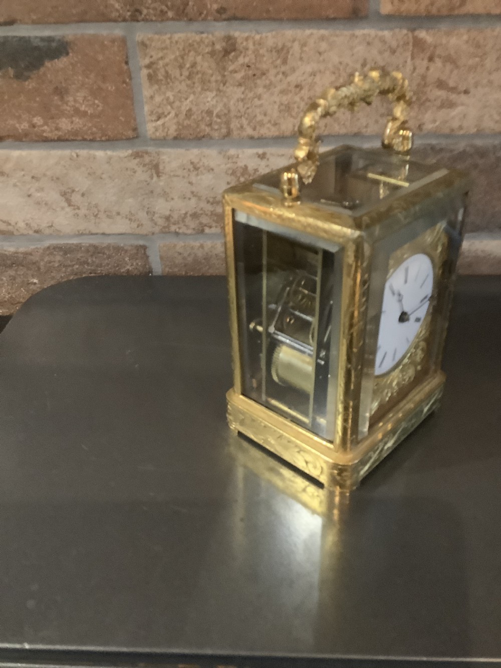 A rectangular gilt brass French carriage clock featuring ornate floral scroll engravings, glass side panels, a decorative carrying handle, and a white enamel dial. View IV