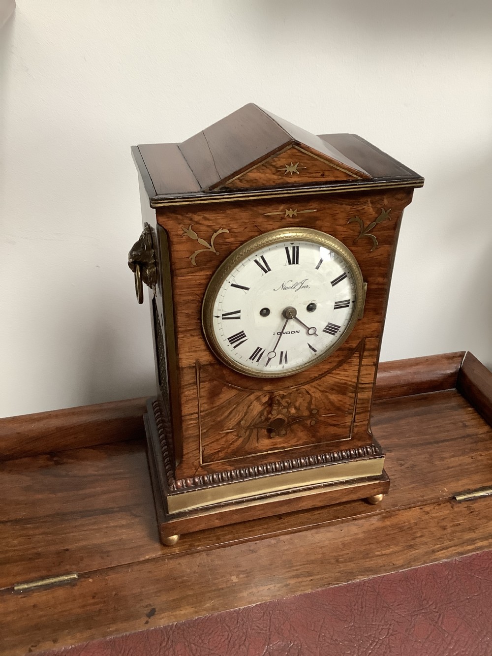 A Regency period wooden table clock with brass inlay, a pediment top, brass lion-head side handles, and a white dial displaying Roman numerals.