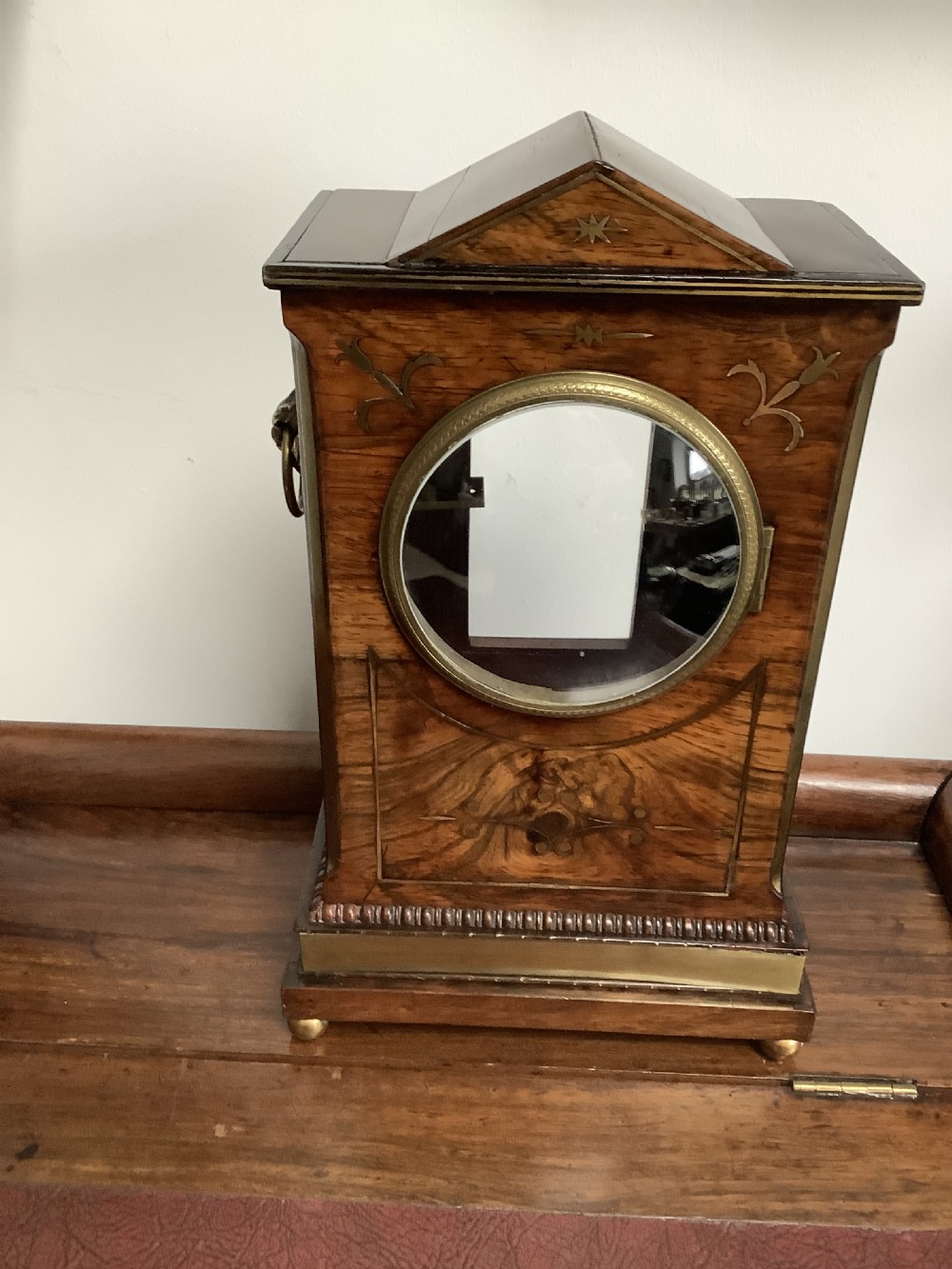 A Regency period wooden table clock with brass inlay, a pediment top, brass lion-head side handles, and a white dial displaying Roman numerals. View XVII