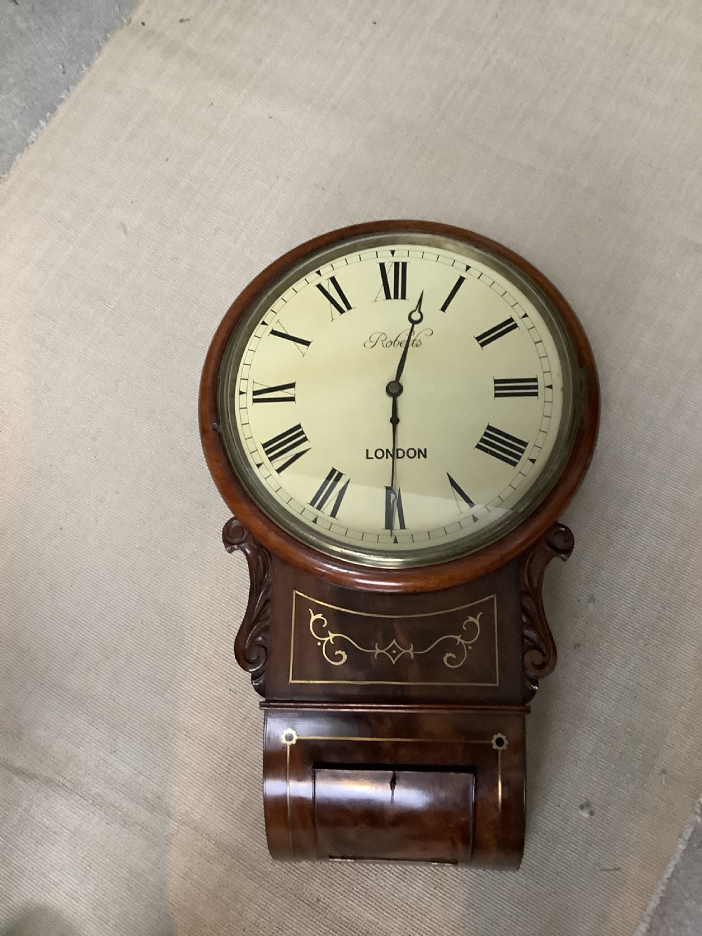 An antique Victorian mahogany drop dial wall clock featuring brass string inlay, carved side ears, and Roman numerals on a cream dial.