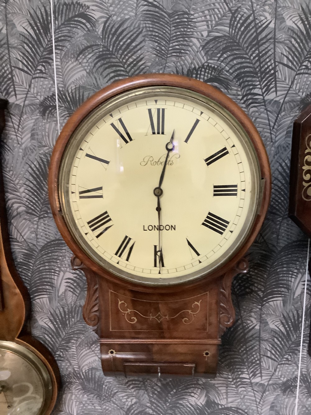 An antique Victorian mahogany drop dial wall clock featuring brass string inlay, carved side ears, and Roman numerals on a cream dial. View III