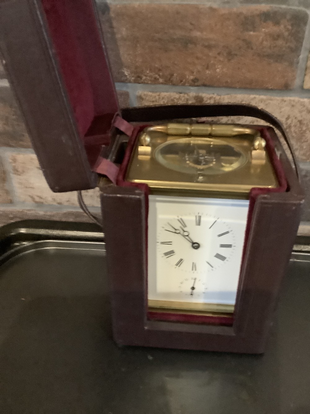 A square dark brown leather travelling case with a front window displaying a white clock dial with black Roman numerals and a small subsidiary dial. View II