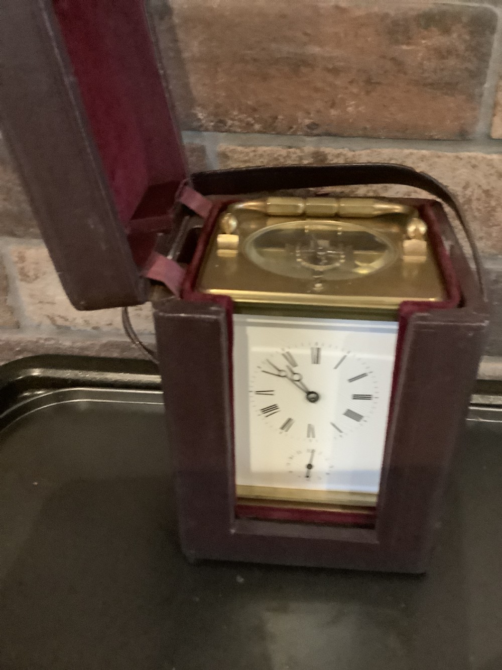 A square dark brown leather travelling case with a front window displaying a white clock dial with black Roman numerals and a small subsidiary dial. View III