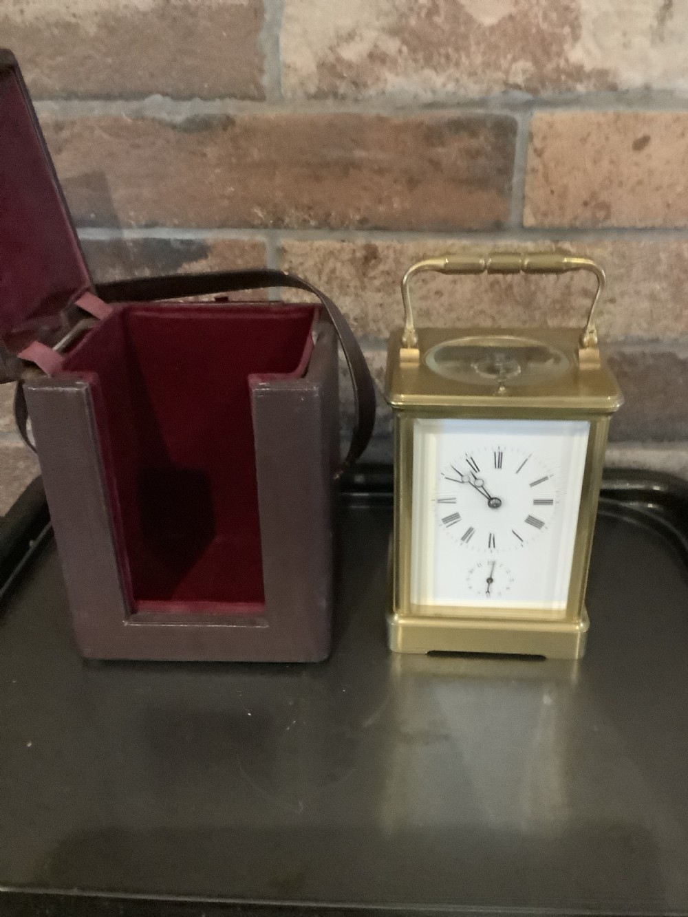 A square dark brown leather travelling case with a front window displaying a white clock dial with black Roman numerals and a small subsidiary dial. View V