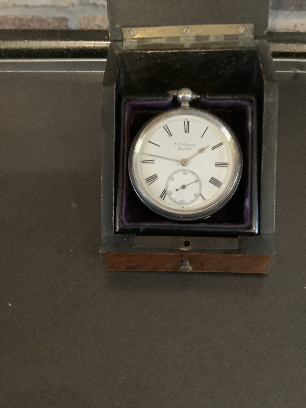 An antique pocket watch with a white enamel dial and Roman numerals displayed open inside a velvet-lined wooden presentation box.