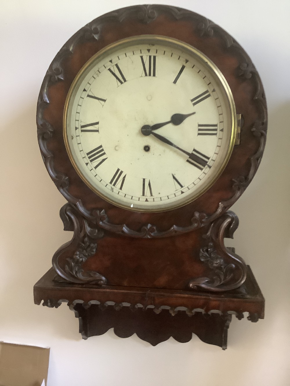 A Victorian carved mahogany fusee wall clock featuring an off-white circular dial with Roman numerals, brass bezel, and ornate matching wooden bracket support.