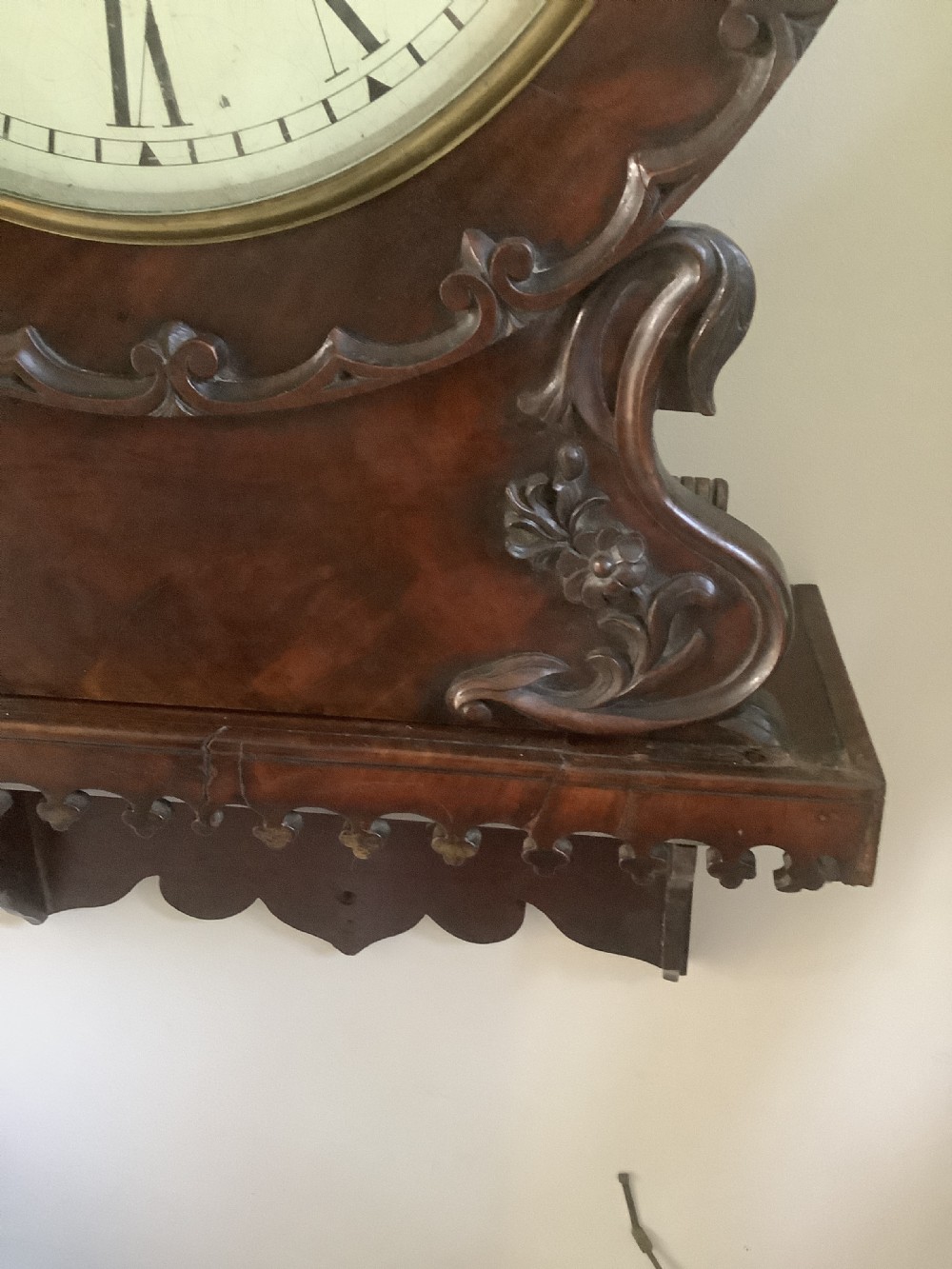 A Victorian carved mahogany fusee wall clock featuring an off-white circular dial with Roman numerals, brass bezel, and ornate matching wooden bracket support. View V