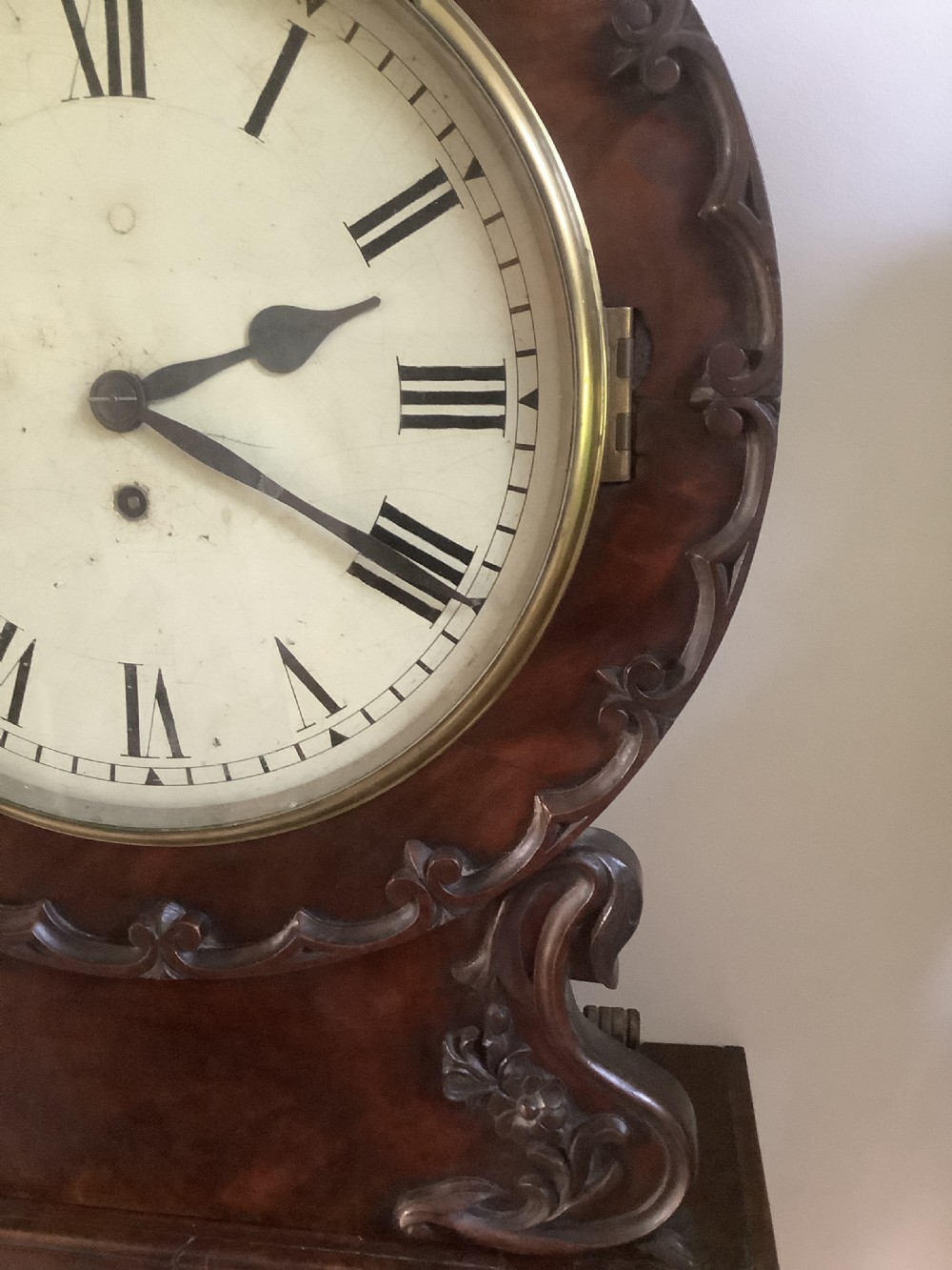 A Victorian carved mahogany fusee wall clock featuring an off-white circular dial with Roman numerals, brass bezel, and ornate matching wooden bracket support. View VI