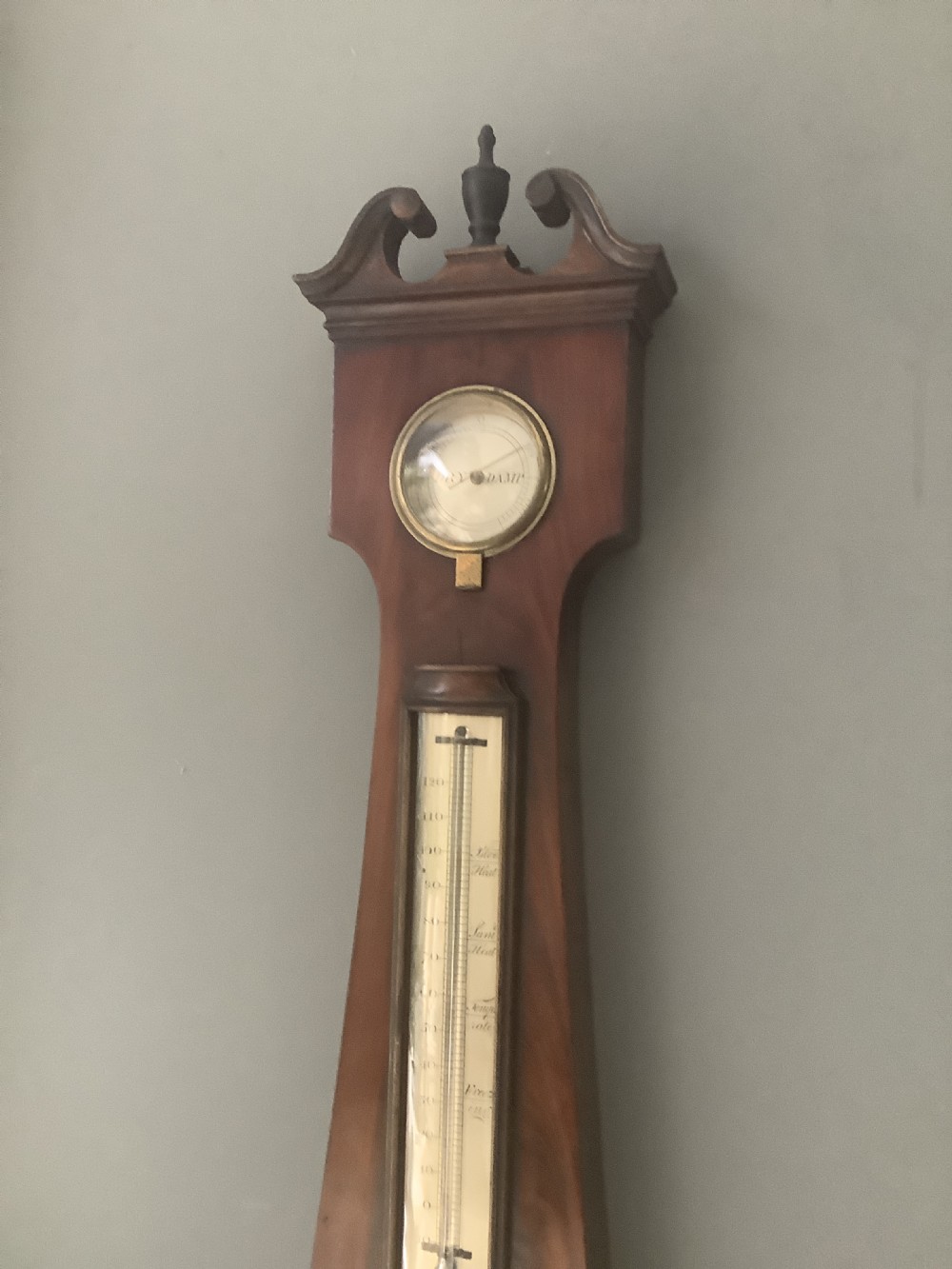 An antique wall-mounted mahogany banjo barometer featuring a swan-neck pediment, a central mercury thermometer tube, and a large circular silvered dial with brass rim. View II