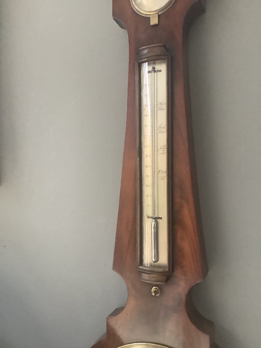 An antique wall-mounted mahogany banjo barometer featuring a swan-neck pediment, a central mercury thermometer tube, and a large circular silvered dial with brass rim. View IV