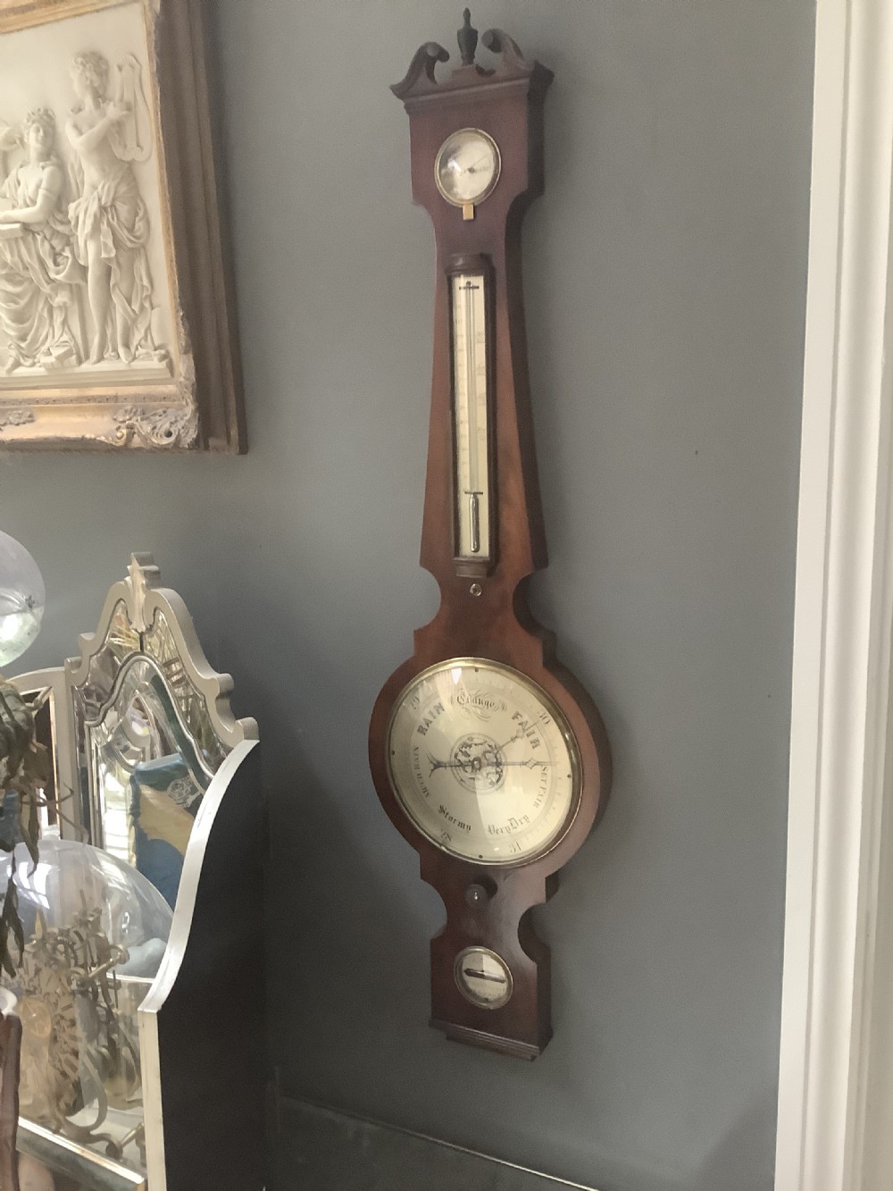An antique wall-mounted mahogany banjo barometer featuring a swan-neck pediment, a central mercury thermometer tube, and a large circular silvered dial with brass rim. View VI