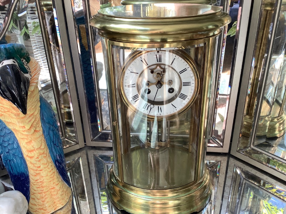 An oval brass four glass mantel clock featuring an enamel dial with Roman numerals and visible internal escapement mechanism.