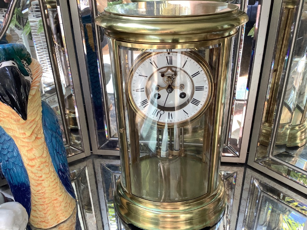 An oval brass four glass mantel clock featuring an enamel dial with Roman numerals and visible internal escapement mechanism. View VI