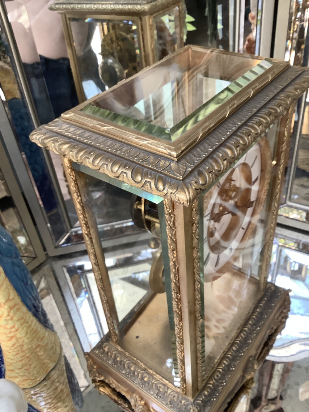 A 19th-century French four-glass mantel clock featuring a gilt brass case with decorative moulding, bevelled glass panels, a white enamel dial with Roman numerals, and a visible mercury pendulum. View VII