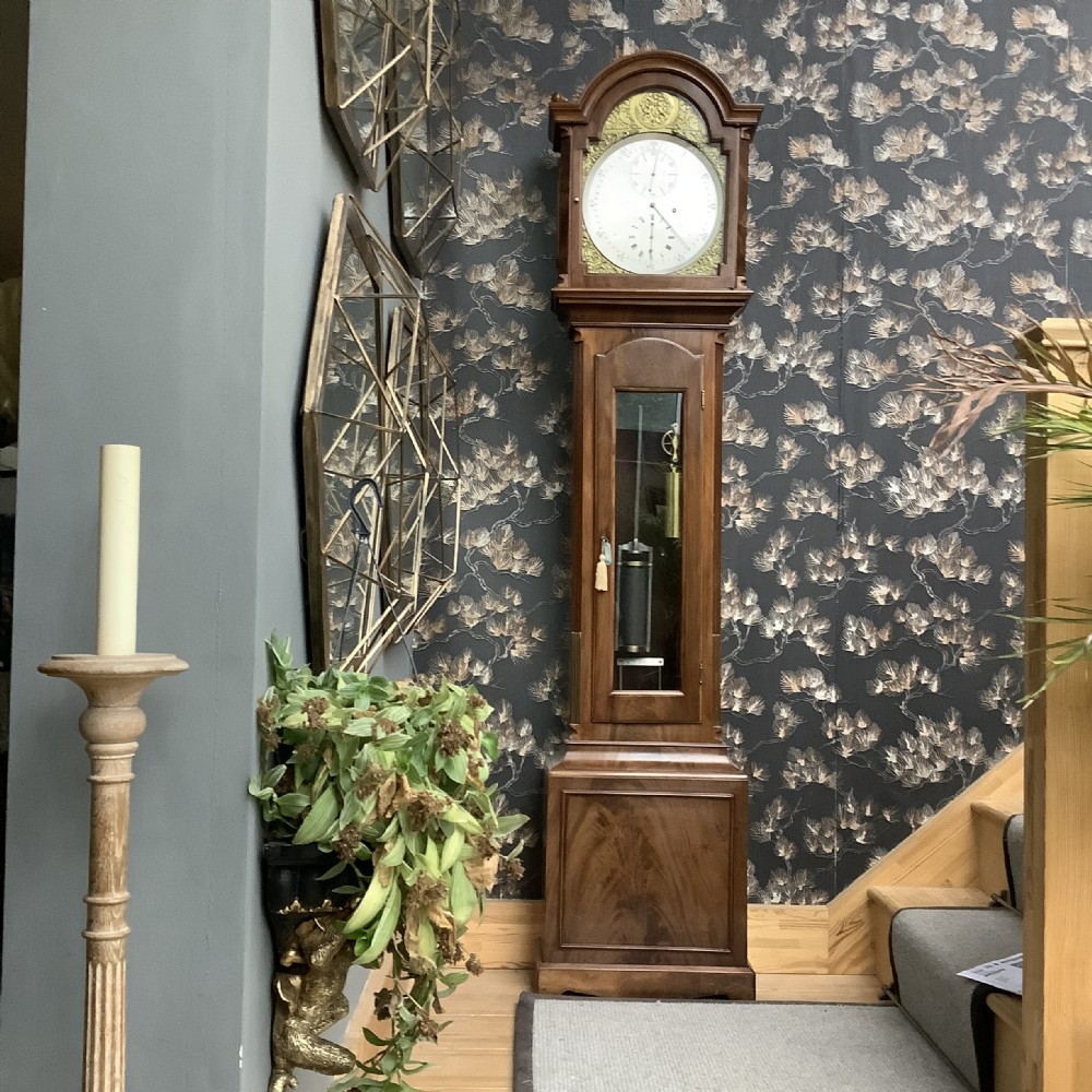 An antique English longcase clock featuring a warm brown timber case, an arched hood, brass spandrels surrounding a clock face, and a central glazed door showing internal weights.