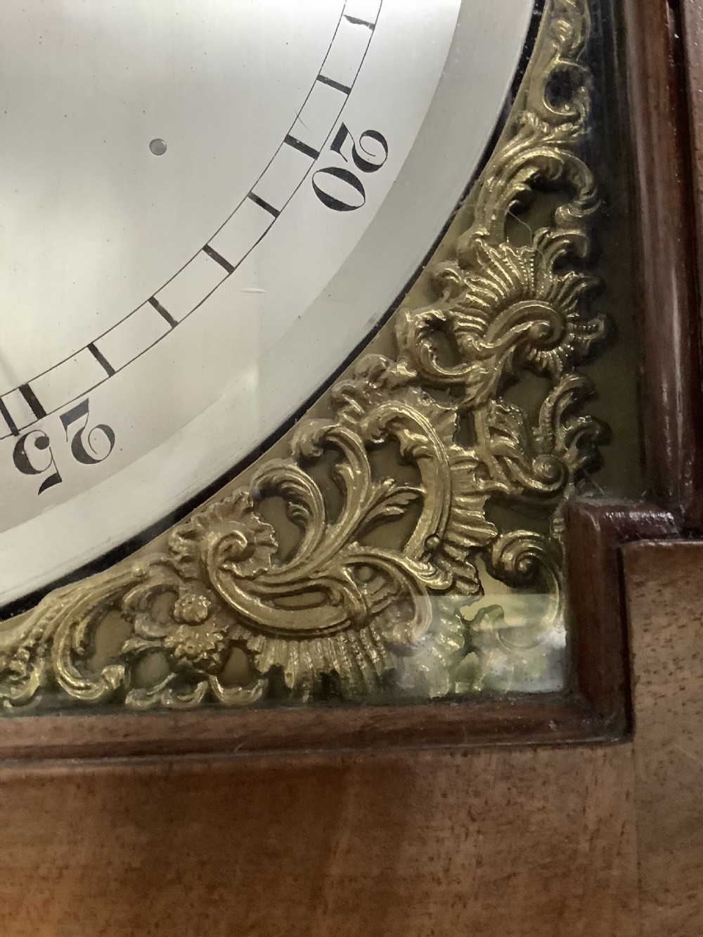 An antique English longcase clock featuring a warm brown timber case, an arched hood, brass spandrels surrounding a clock face, and a central glazed door showing internal weights. View III