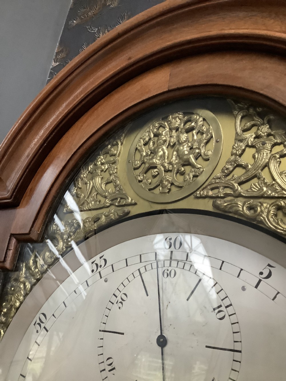 An antique English longcase clock featuring a warm brown timber case, an arched hood, brass spandrels surrounding a clock face, and a central glazed door showing internal weights. View VI