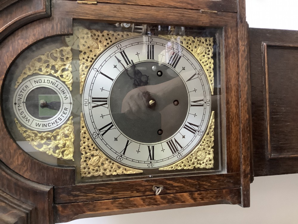 A dark oak freestanding grandmother clock with a pointed arched hood, glass door, and a brass dial featuring chime selection sub-dials. View III