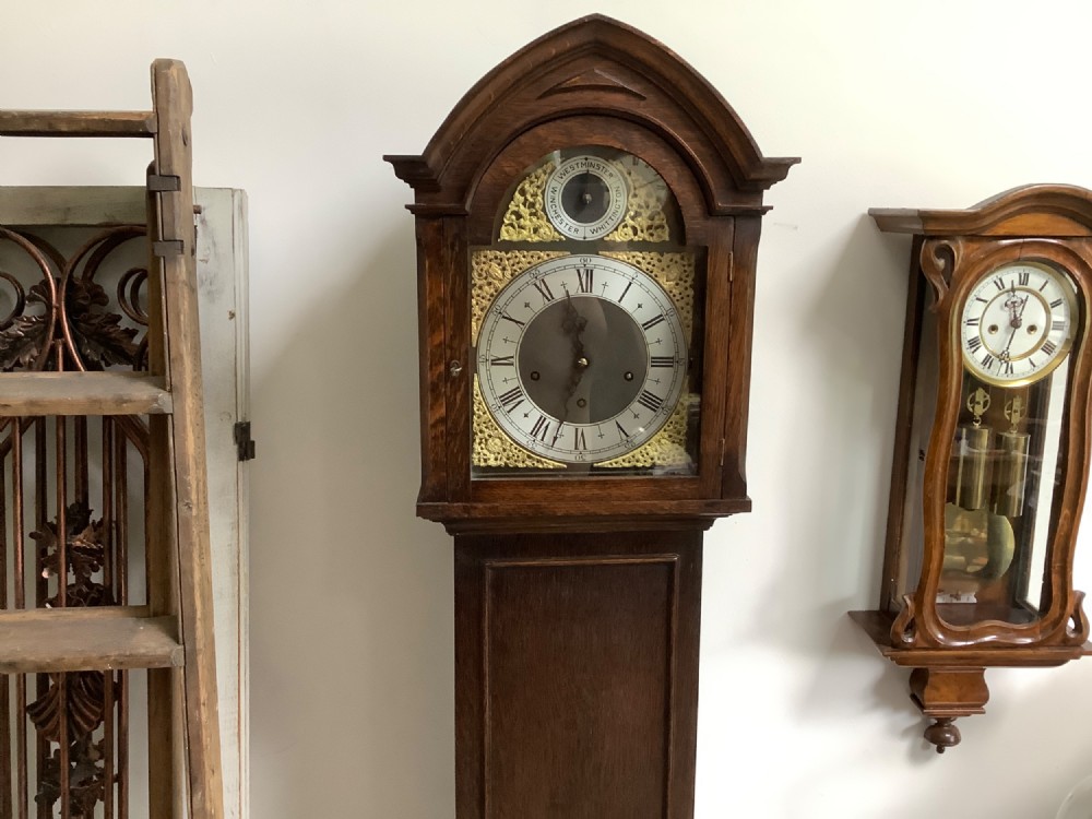 A dark oak freestanding grandmother clock with a pointed arched hood, glass door, and a brass dial featuring chime selection sub-dials. View II