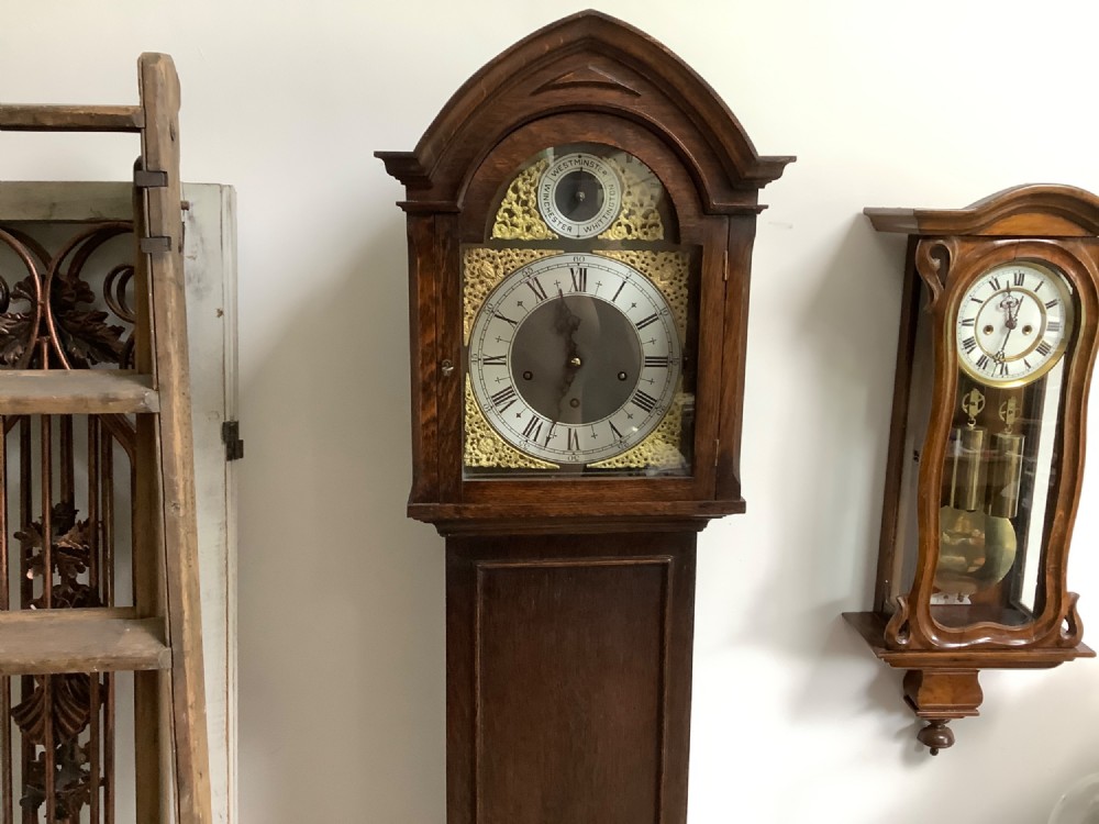 A dark oak freestanding grandmother clock with a pointed arched hood, glass door, and a brass dial featuring chime selection sub-dials. View V