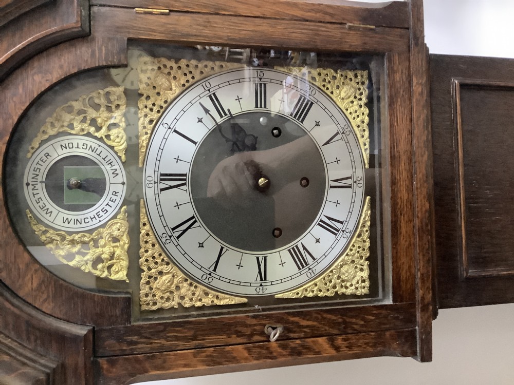 A dark oak freestanding grandmother clock with a pointed arched hood, glass door, and a brass dial featuring chime selection sub-dials. View VI