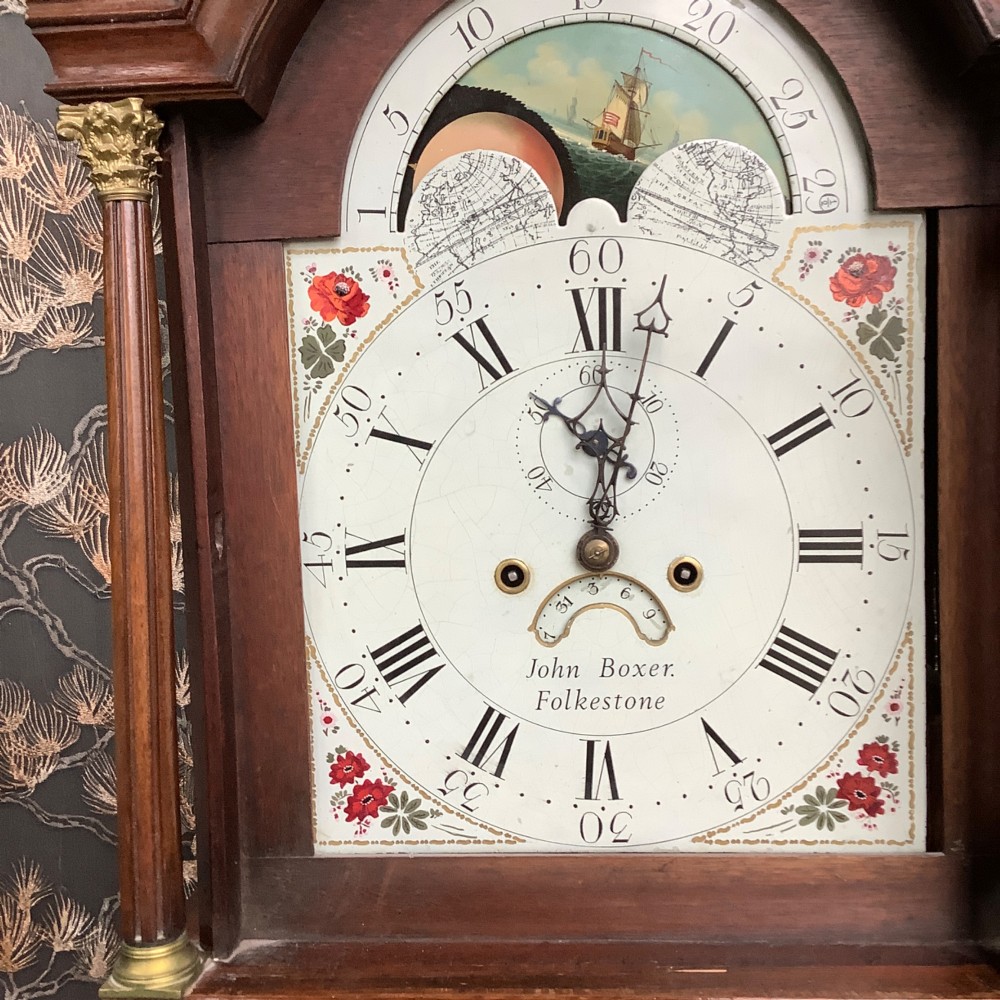 An antique mahogany grandfather clock featuring a painted dial with Roman numerals, a swan-neck pediment, reeded columns, and a polished timber trunk door. View III