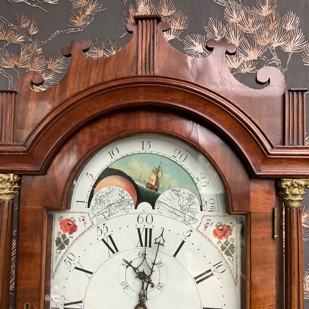 An antique mahogany grandfather clock featuring a painted dial with Roman numerals, a swan-neck pediment, reeded columns, and a polished timber trunk door. View IV